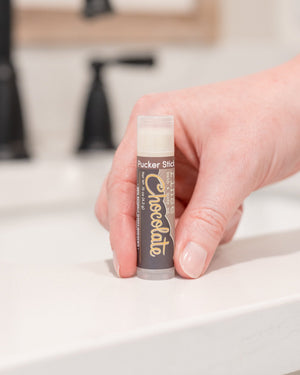 Chocolate Pucker Stick lip balm in hand, cap off — a pocket-sized twist-up balm that smells like real cocoa. Rinse Bath & Body Co.