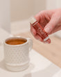 A hand holding the Chai Tea Latte lip balm beside a mug of spiced milk tea — cinnamon, cardamom and vanilla for soft winter lips. Rinse Bath & Body Co.