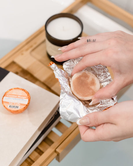 Two hands unwrapping the foil from a Coconut Mango Tub Truffle over a bamboo bath caddy, a lit candle beside them — a hot bath that smells tropical. Rinse Bath & Body Co.