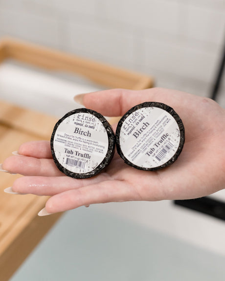 An open palm holding two black-labelled Birch Tub Truffles above a filled bath with a bamboo caddy behind — woodsy and clean, like a cabin. Rinse Bath & Body Co.