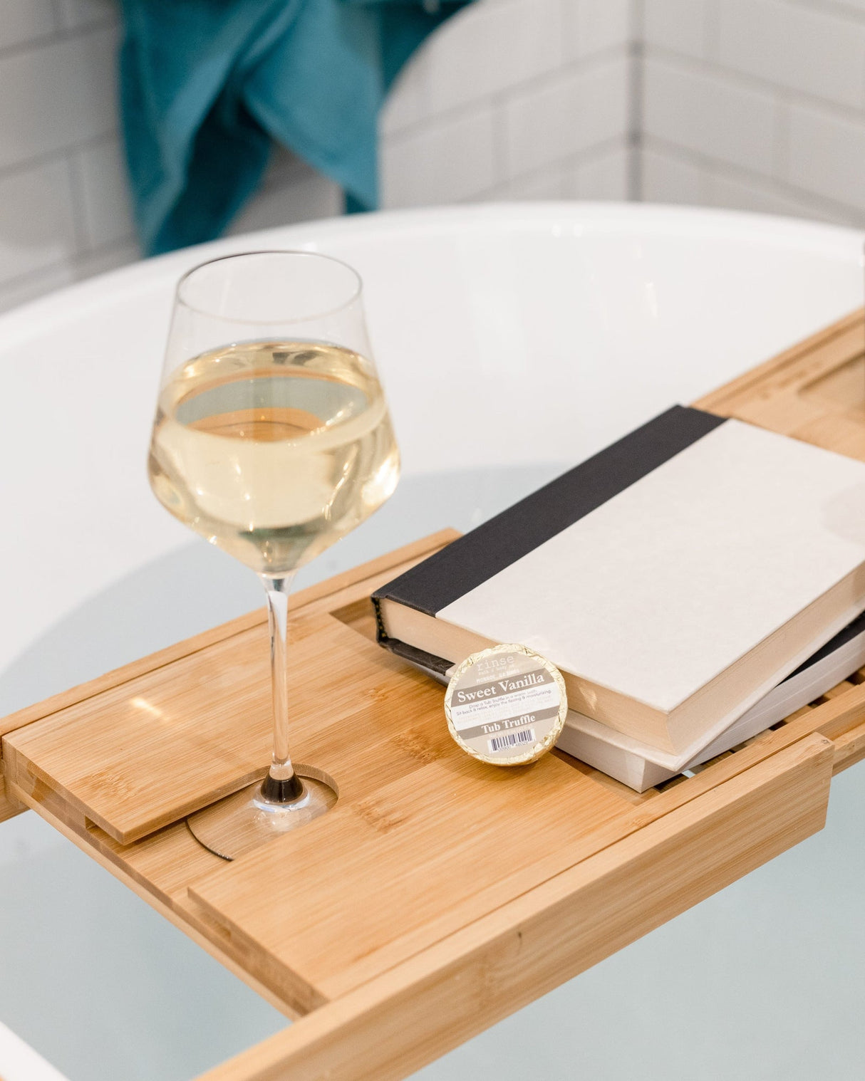 A Sweet Vanilla Tub Truffle on a bamboo bath caddy with a glass of white wine, a book and a candle, beside a filled freestanding tub. Rinse Bath & Body Co.