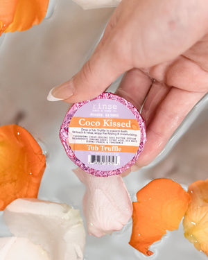 A hand holding a Coco Kissed Tub Truffle above a bath scattered with orange flower petals — cocoa on cocoa, skin doesn't stand a chance. Rinse Bath & Body Co.