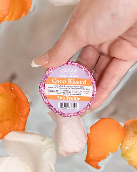 A hand holding a Coco Kissed Tub Truffle above a bath scattered with orange flower petals — cocoa on cocoa, skin doesn't stand a chance. Rinse Bath & Body Co.