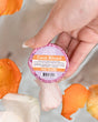 A hand holding a Coco Kissed Tub Truffle above a bath scattered with orange flower petals — cocoa on cocoa, skin doesn't stand a chance. Rinse Bath & Body Co.