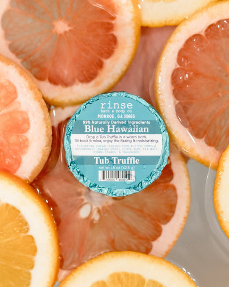 A teal-labelled Blue Hawaiian Tub Truffle resting among floating grapefruit and orange slices in bath water — blue water, citrus, no flight required. Rinse Bath & Body Co.
