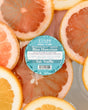 A teal-labelled Blue Hawaiian Tub Truffle resting among floating grapefruit and orange slices in bath water — blue water, citrus, no flight required. Rinse Bath & Body Co.