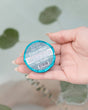 An open palm holding a teal foil-wrapped Euca-Mint Tub Truffle above a filled bath, eucalyptus stems trailing behind. Rinse Bath & Body Co.