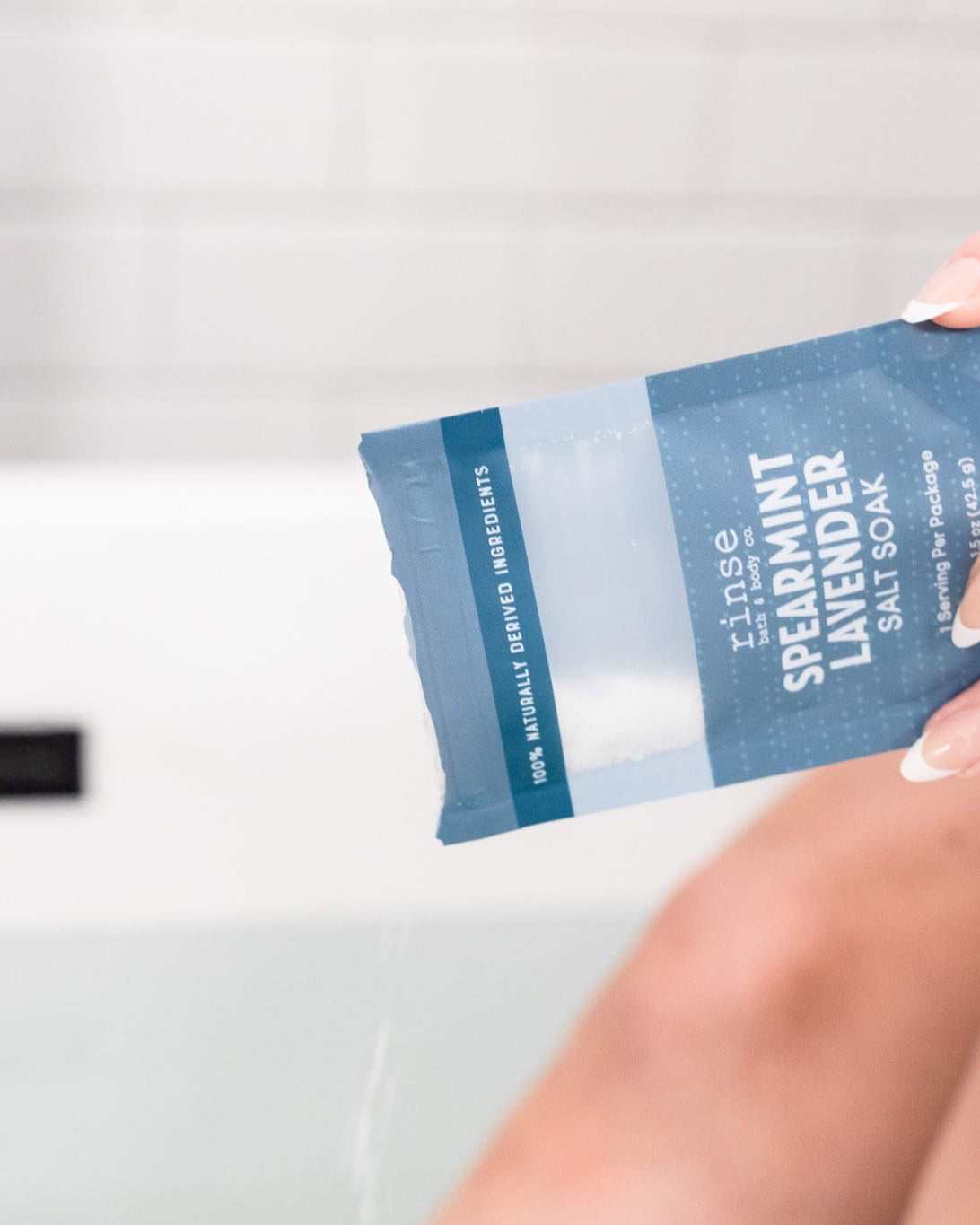 Hand pouring Spearmint Lavender Salt Soak into a filled tub against white subway tile — cool spearmint and soft lavender for a quiet soak