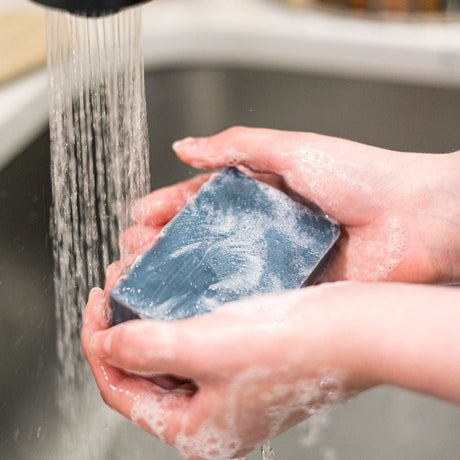 Hands lathering the marbled navy Flannel bar under running water — woodsmoke, cedar and vanilla in a cocoa-butter base. Rinse Bath & Body Co.