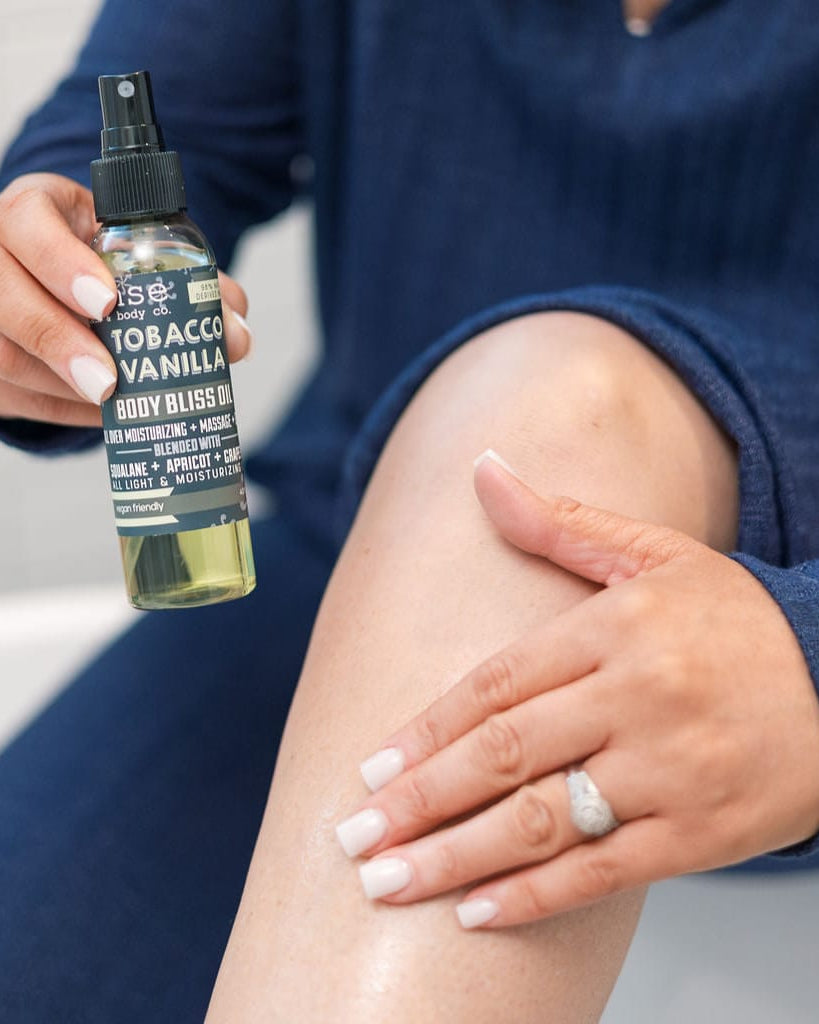 Tobacco Vanilla Body Bliss oil in use, smoothed into skin — dark tobacco leaf over warm vanilla in a fast-absorbing oil. Rinse Bath & Body Co.