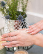 Hands working granular sugar scrub over the back of a hand beside a silver mesh planter of rosemary and blue thistle, an open jar at the edge of frame — the scrub step, before it rinses to a lotion finish. Rinse Bath & Body Co.