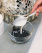 A spoonful of mask paste poured into a glass bowl of black charcoal powder, mixing CharClay Face Mask by hand. Rinse Bath & Body Co.