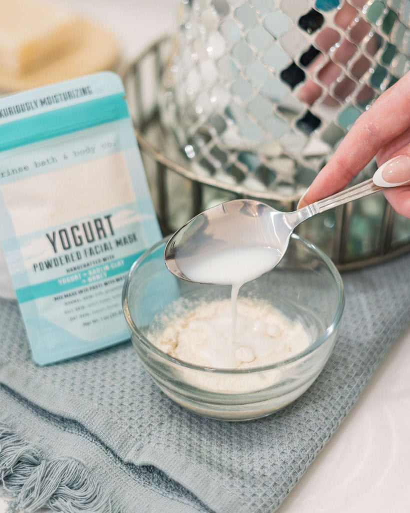 A spoonful of Yogurt Face Mask powder lifted from a bowl, the pouch resting on grey linen beside it. Rinse Bath & Body Co.