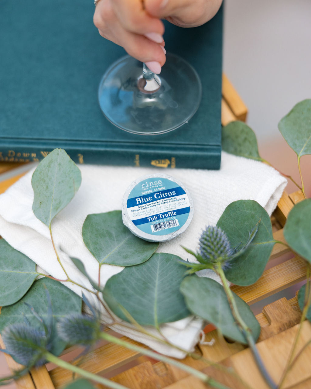 A hand lifting a wine glass over a wooden bath tray holding a Blue Citrus Tub Truffle, eucalyptus, thistle and a cloth-bound book — three bright baths, boxed. Rinse Bath & Body Co.