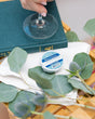 A Blue Citrus Tub Truffle on a folded white cloth with eucalyptus and thistle on a wooden bath tray, a hand lifting a wine glass above it — bright citrus for a slow soak. Rinse Bath & Body Co.
