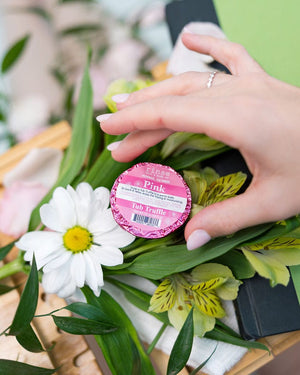 A hand setting a pink foil-wrapped Tub Truffle among white daisies and alstroemeria on a wooden tray — cocoa butter and unapologetic pink. Rinse Bath & Body Co.