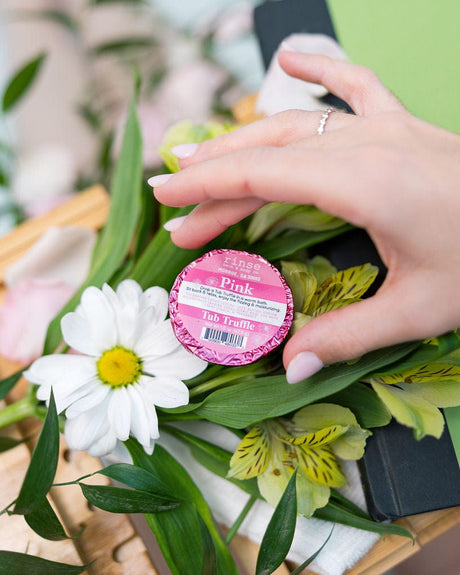 A hand setting a pink foil-wrapped Tub Truffle among white daisies and alstroemeria on a wooden tray — cocoa butter and unapologetic pink. Rinse Bath & Body Co.