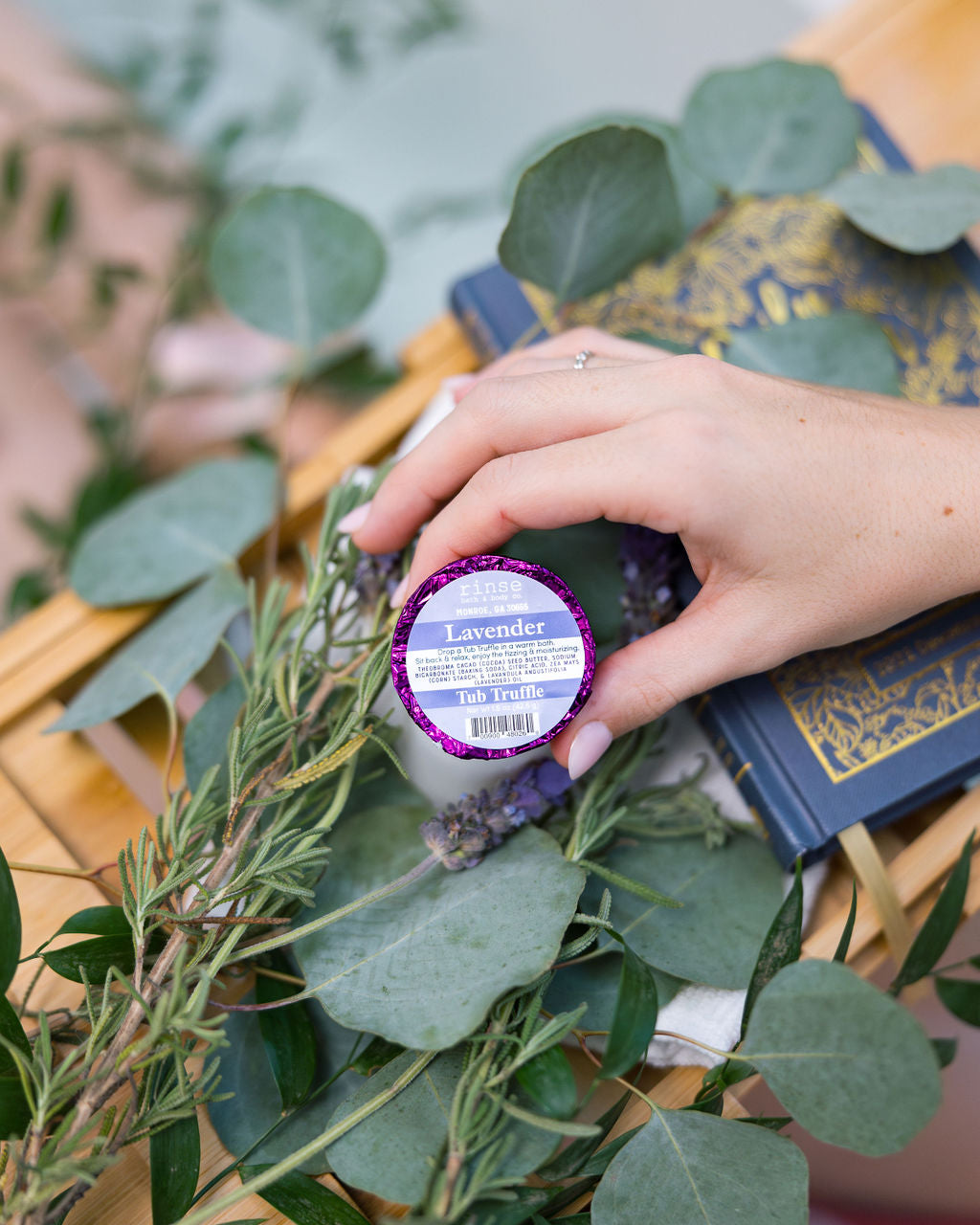 A hand placing a purple foil-wrapped Lavender Tub Truffle among fresh lavender and eucalyptus on a wooden bath tray — three lavender baths, boxed. Rinse Bath & Body Co.