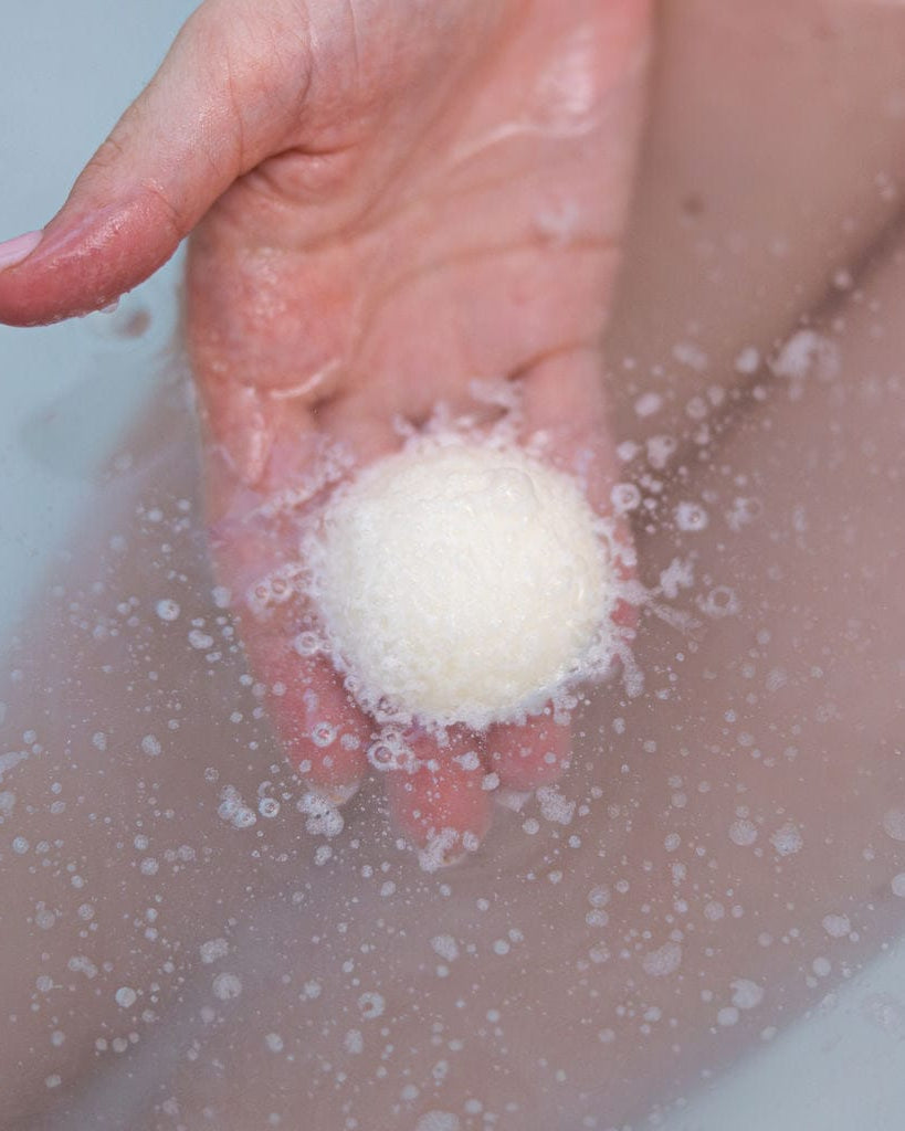 A hand holding a white Flannel Tub Truffle just under the surface of a warm bath, fizzing into the water — cocoa butter softening as it dissolves. Rinse Bath & Body Co.