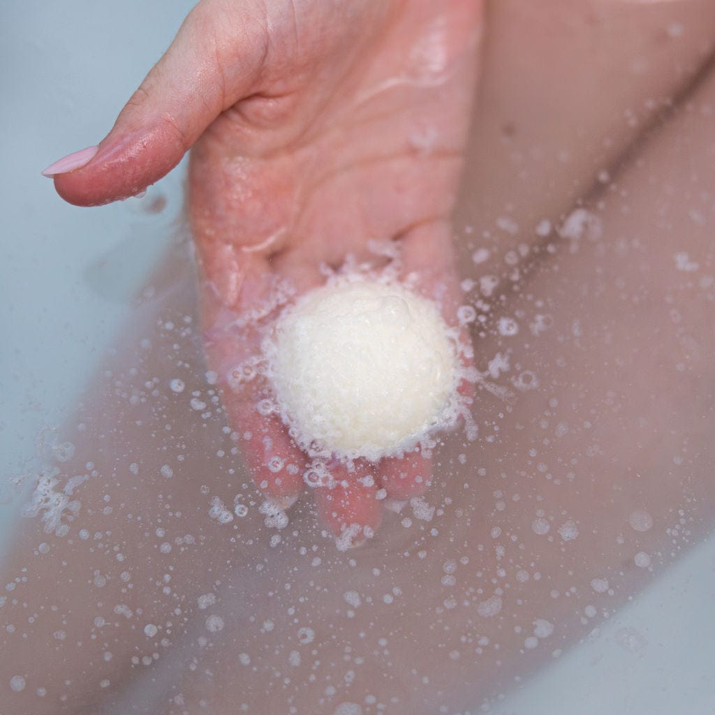 A hand holding a white cocoa butter Tub Truffle just under the surface of a warm bath, fizzing as it dissolves and releasing the cocoa butter into the water. Rinse Bath & Body Co.