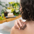 Seen from behind, a woman rubs the textured brown Body Conditioning Polish bar across her wet shoulder, a bath tray and greenery blurred behind — a solid exfoliating scrub bar worked straight onto damp skin. Rinse Bath & Body Co.