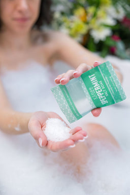 Hand pouring sparkling Speppermint salts into a palm above bubbly bathwater — floral bouquet soft behind