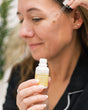 A woman holding the Pure Marula Oil bottle beside her face, outdoors in soft daylight. Rinse Bath & Body Co.
