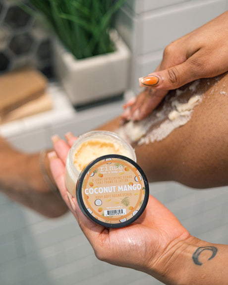 A person applies a white exfoliating scrub to their leg while holding a jar of Rinse Coconut Mango body scrub in their other hand, set against a blurred bathroom background.