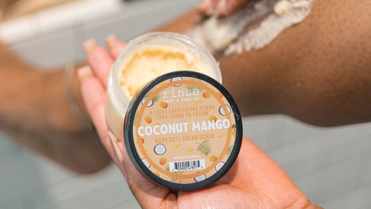 A person applies a white exfoliating scrub to their leg while holding a jar of Rinse Coconut Mango body scrub in their other hand, set against a blurred bathroom background.