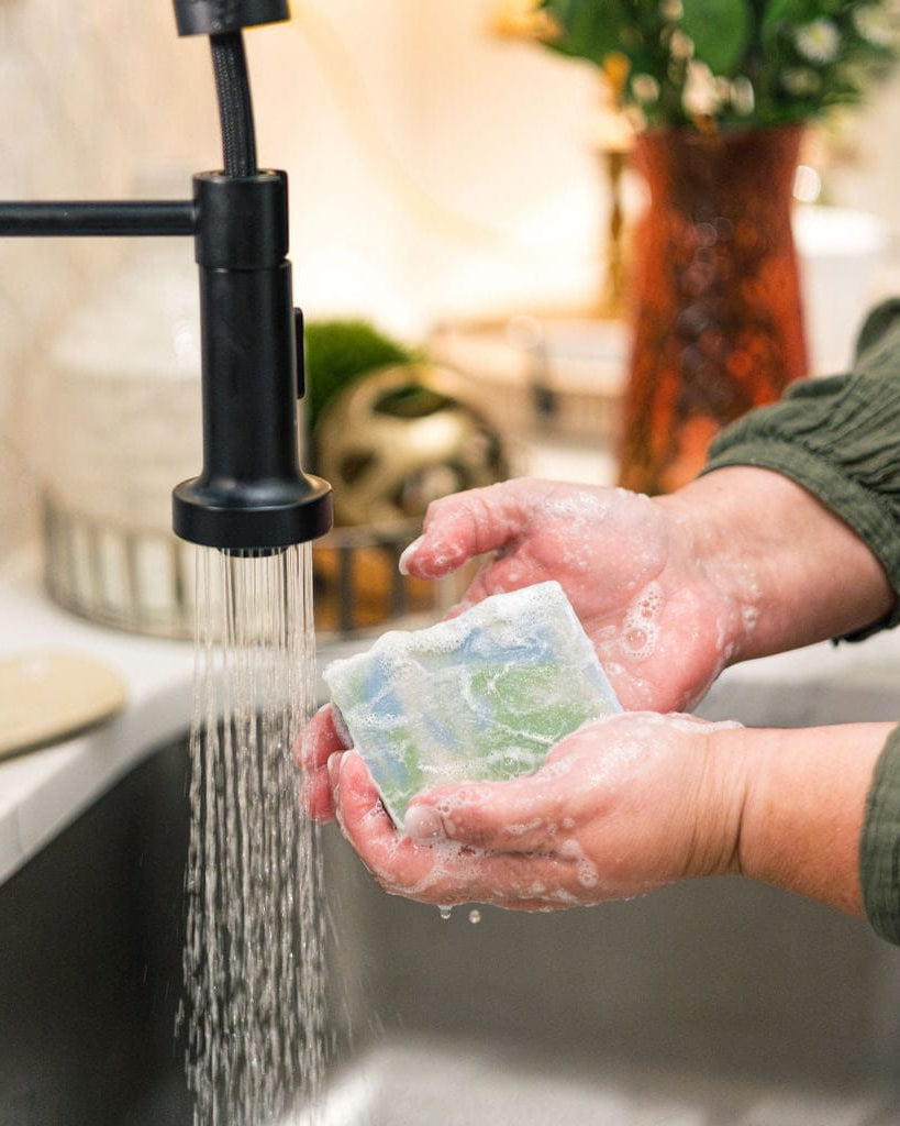 Hands lathering Rainforest under a black faucet — stainless sink marble counter and orange vase soft behind