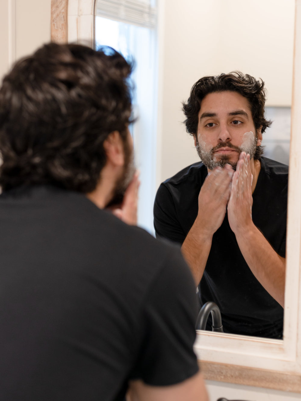 Man massaging thick white lather into his dark beard — bathroom mirror catching soft morning light