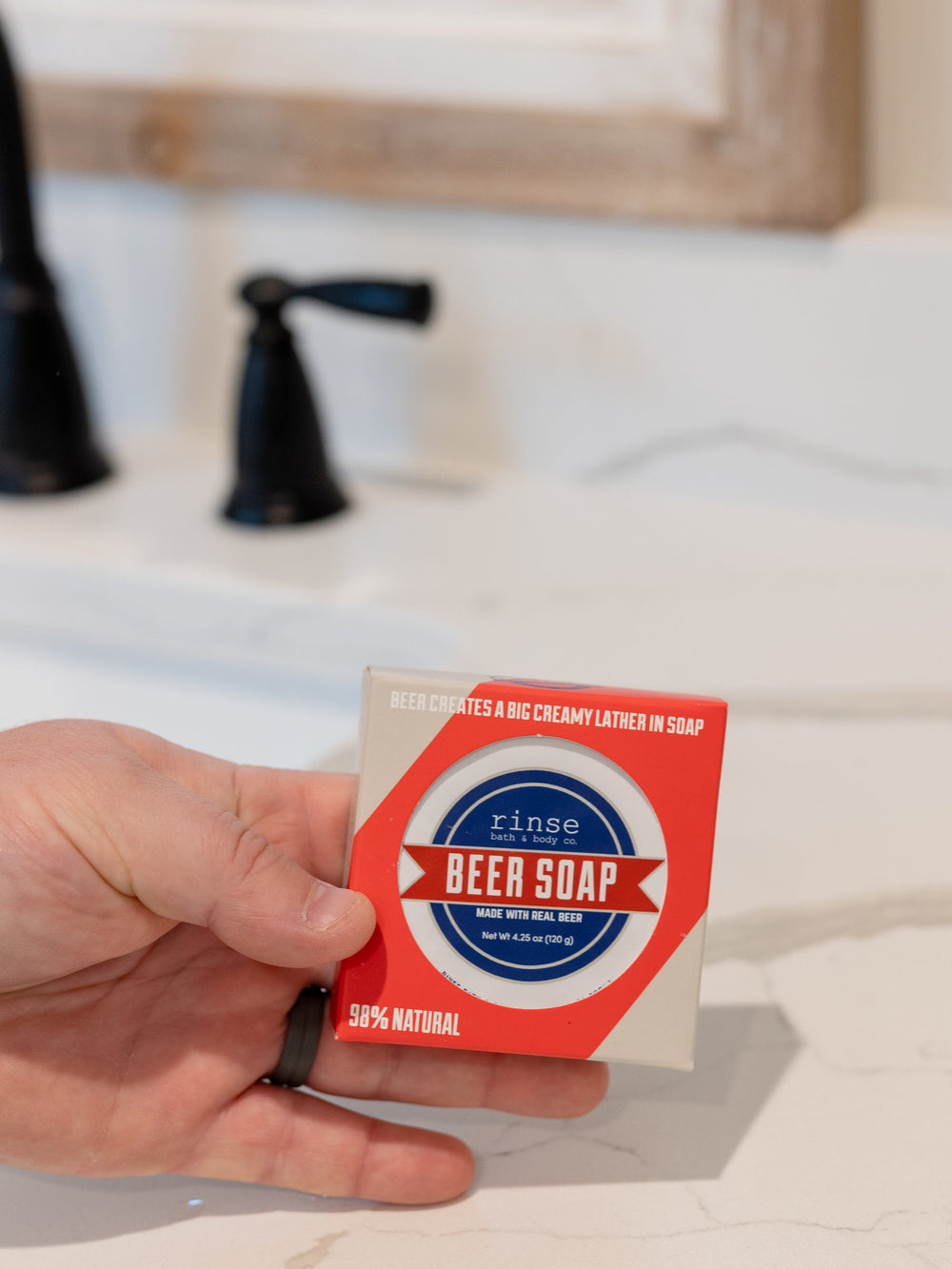 Hand holding Blue Ribbon Beer Soap carton over marble sink — black faucet soft behind in bright bathroom light