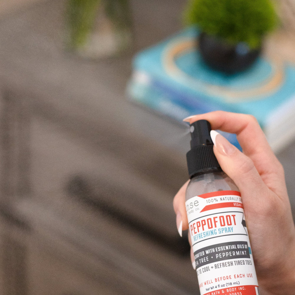 A hand holding Peppofoot Refreshing Spray beside a bedside table with a plant and stacked books — the last step before bed for tired feet. Rinse Bath & Body Co.
