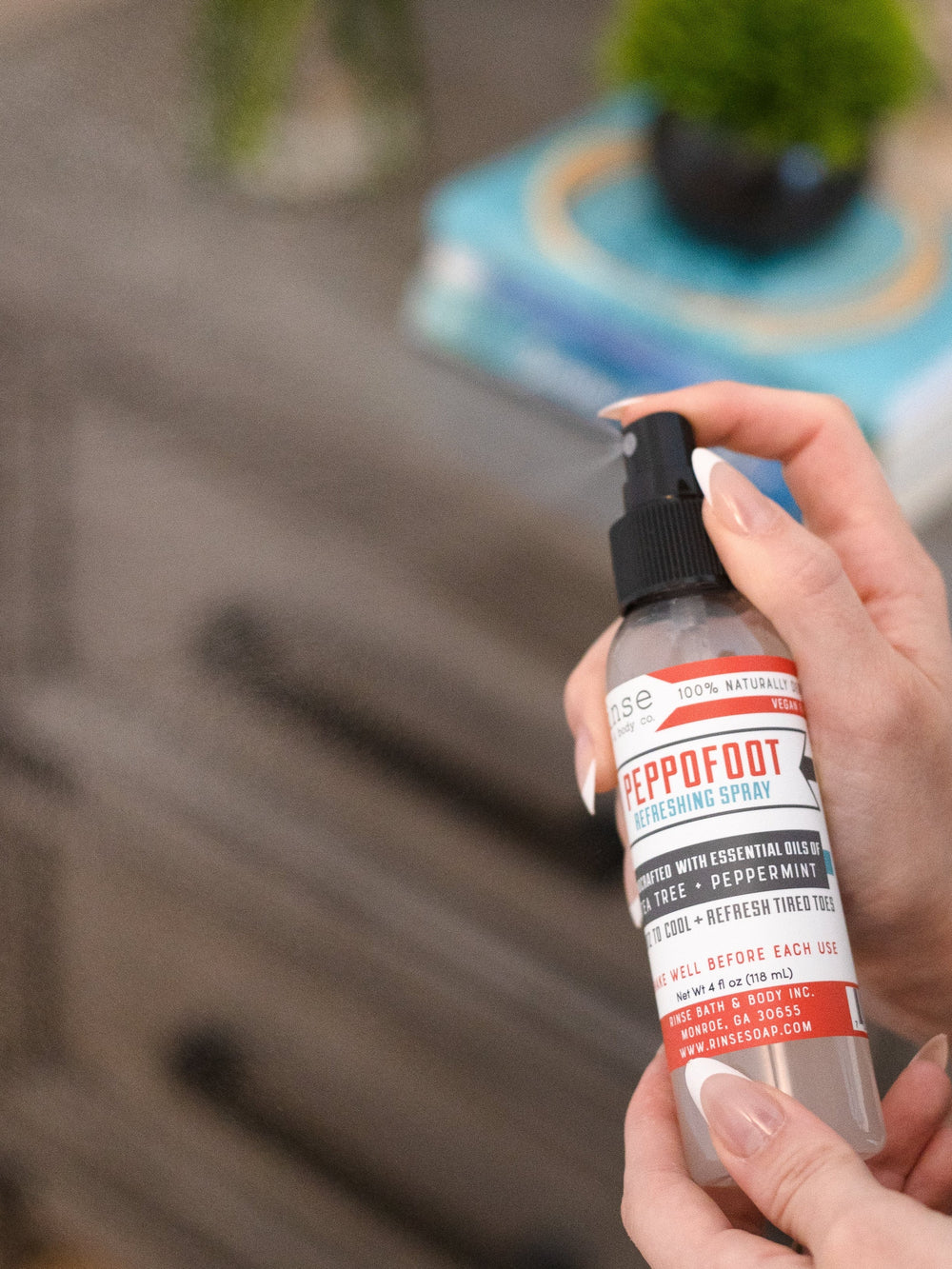 A hand holding Peppofoot Refreshing Spray beside a bedside table with a plant and stacked books — the last step before bed for tired feet. Rinse Bath & Body Co.