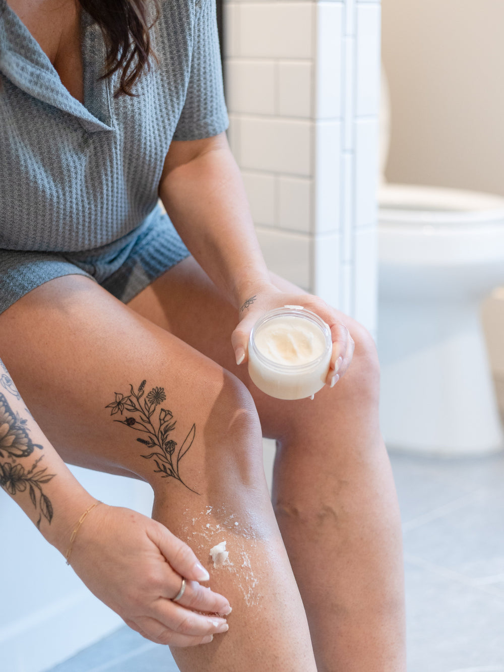 A woman in a grey waffle robe sits on the edge of a white bathtub, an open jar of Body Buff sugar scrub in one hand and a smear of it worked into her shin, subway tile behind — the scrub step of the Scrub & Shine ritual. Rinse Bath & Body Co.