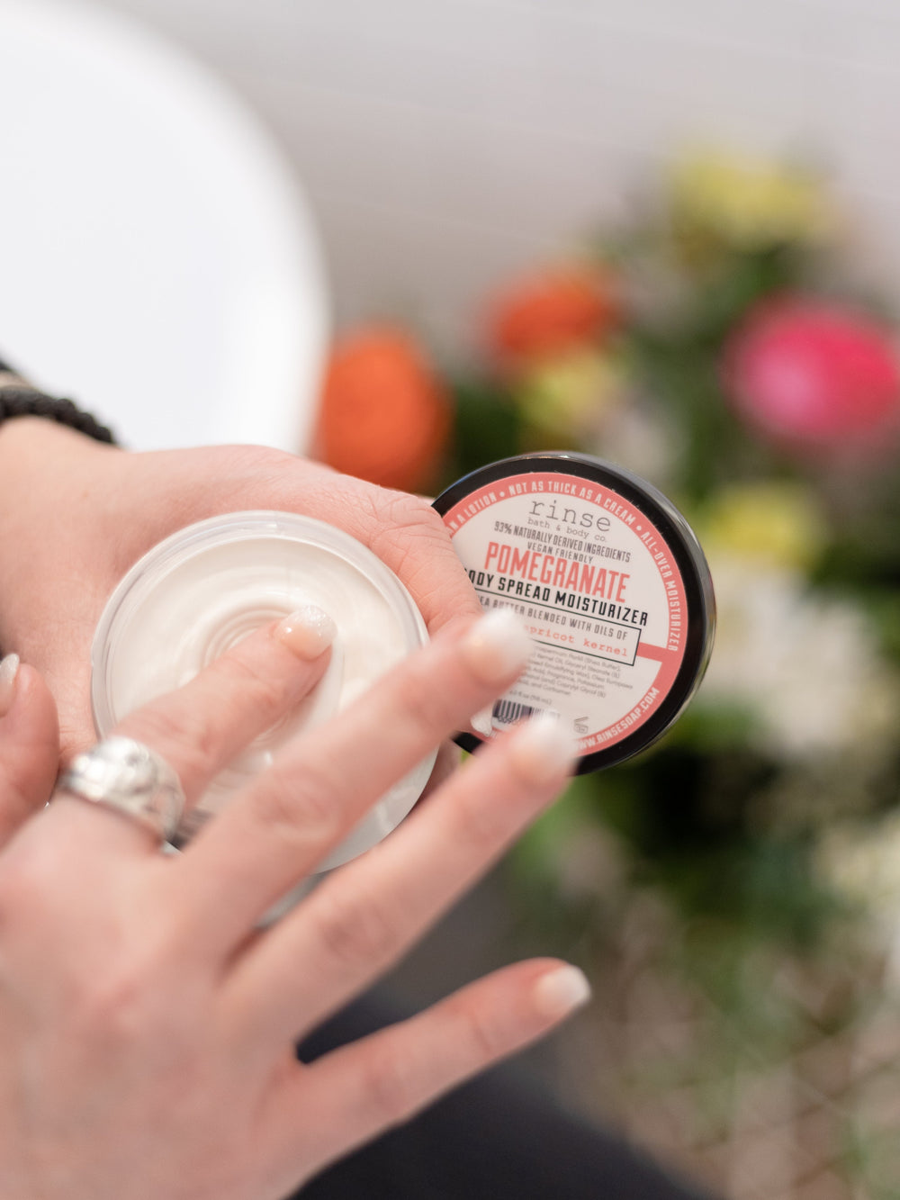 Finger dipping into Pomegranate Body Spread jar — silver ring and soft pink-yellow flower bokeh behind
