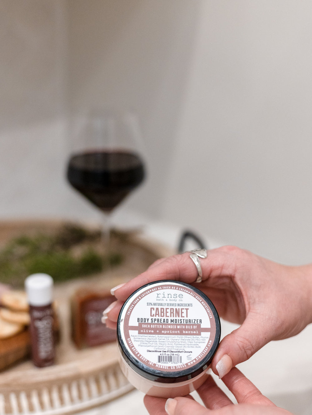 Hands presenting Cabernet Body Spread jar — red wine and wooden snack tray softly glowing behind
