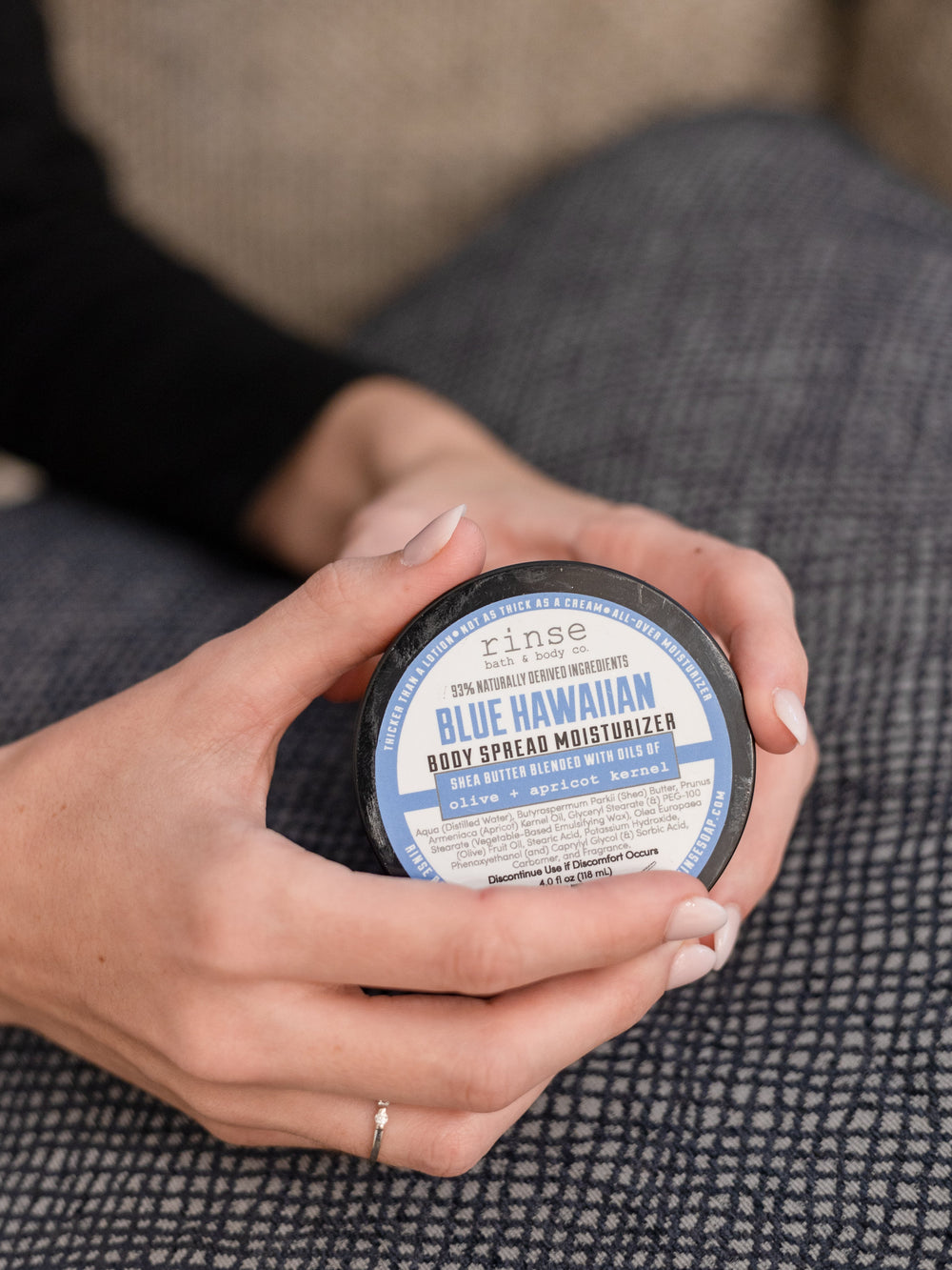 Hands cupping closed Blue Hawaiian Body Spread jar — blue label and black lid on soft gray grid fabric