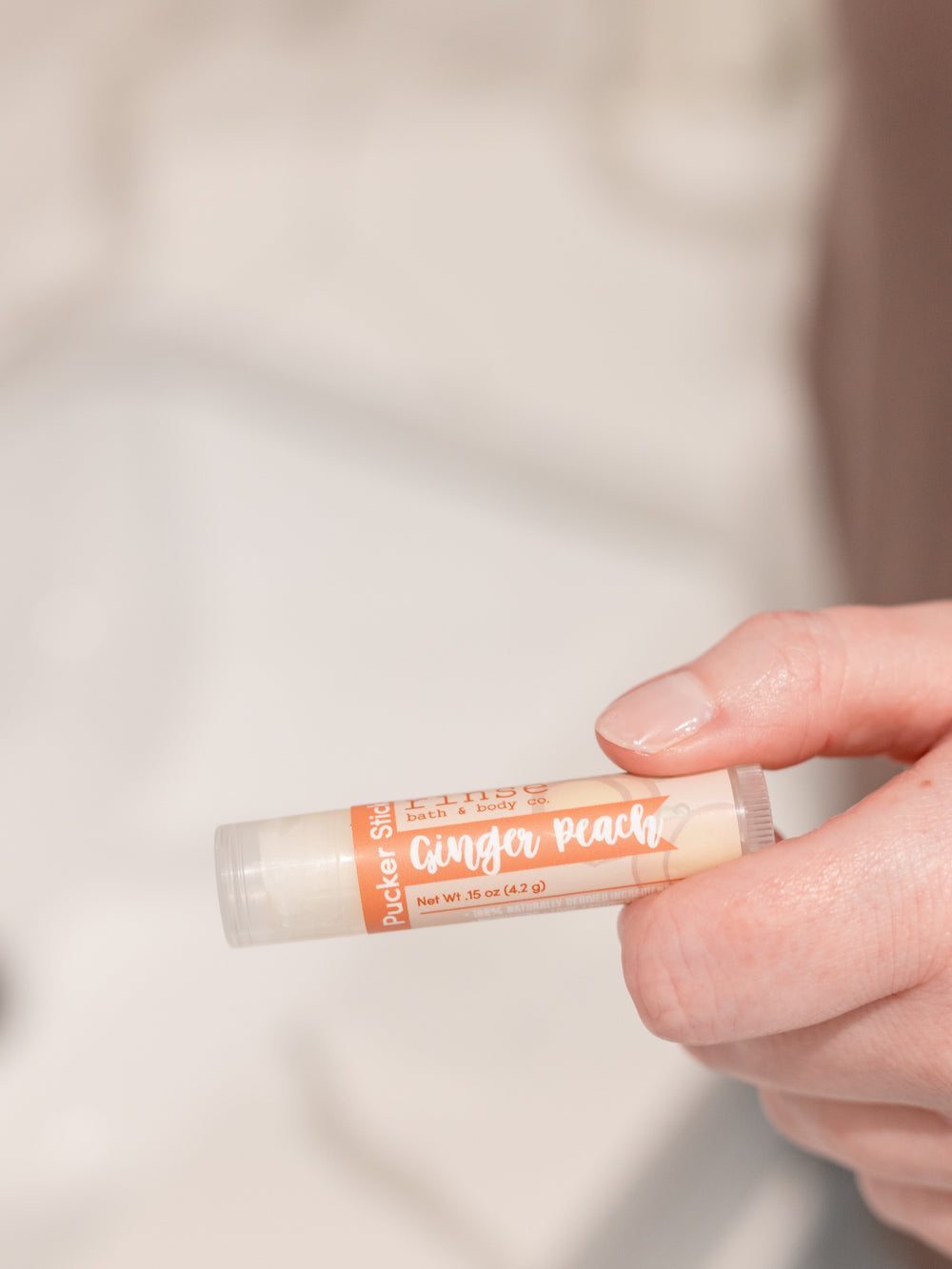 Hand holding Ginger Peach Pucker Stick horizontally — orange cream label in soft bathroom bokeh