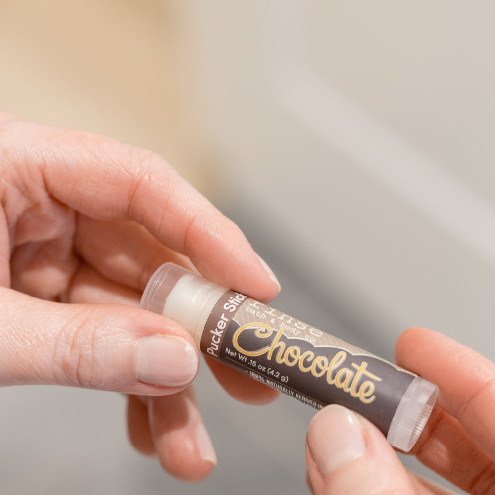 Chocolate lip balm in use, the tube held up to the light — cocoa butter and shea for dry, chapped lips. Rinse Bath & Body Co.