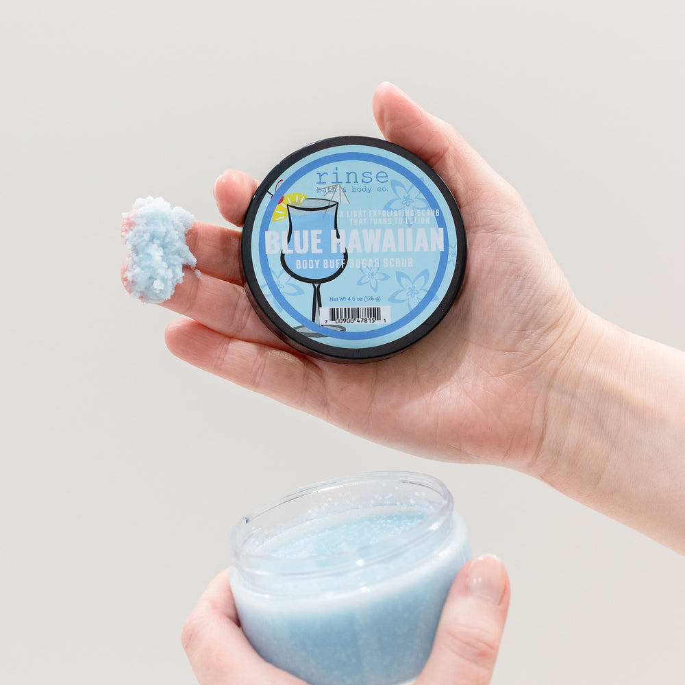 Two hands holding a jar of light blue scrub and a round tin of the same product against a plain light gray background. The round tin label reads 'tins' and 'BLUE HAWAIIAN'.