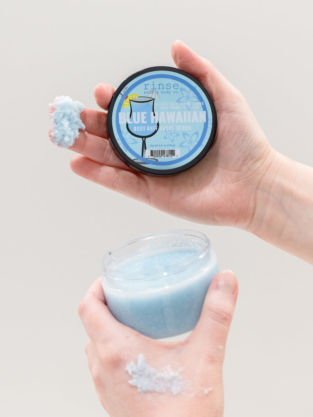 Hands holding Blue Hawaiian scrub with a dollop on the lid — sparkly blue sugar paste on light gray