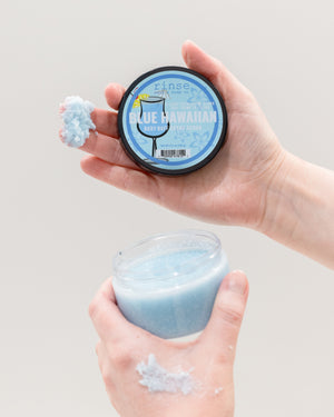 Two hands holding a jar of light blue scrub and a round tin of the same product against a plain light gray background. The round tin label reads 'tins' and 'BLUE HAWAIIAN'.