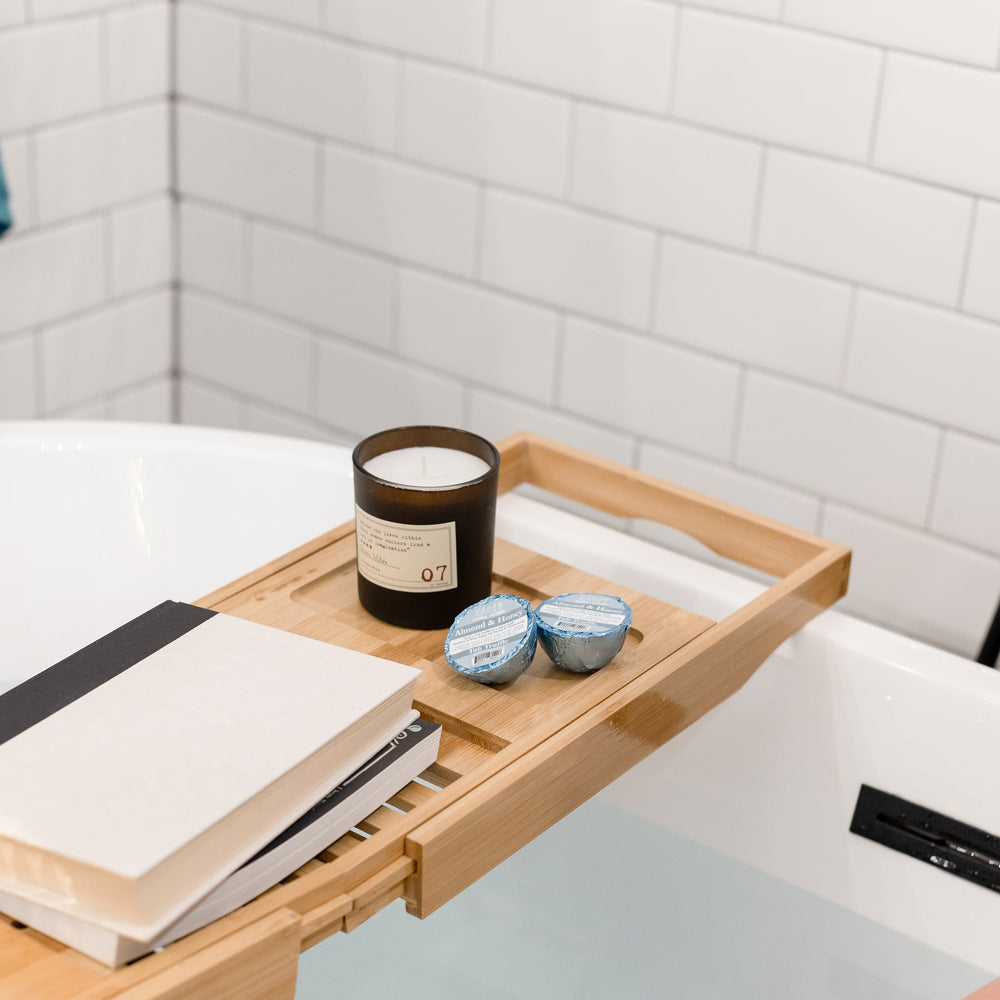 A bamboo caddy laid across a filled tub with a candle, a book and two Almond Honey Tub Truffles, bare legs in the water below — three almond honey baths, boxed. Rinse Bath & Body Co.