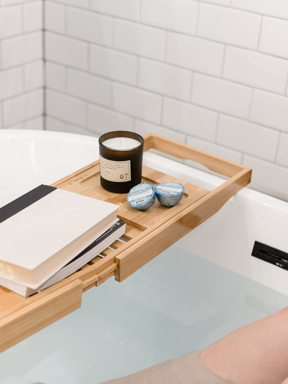 A bamboo caddy laid across a filled tub with a candle, a book and two Almond Honey Tub Truffles, bare legs in the water below — three almond honey baths, boxed. Rinse Bath & Body Co.