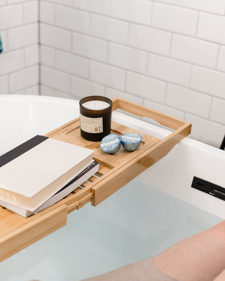 A bamboo caddy laid across a filled tub with a candle, a book and two Almond Honey Tub Truffles, bare legs in the water below — three almond honey baths, boxed. Rinse Bath & Body Co.