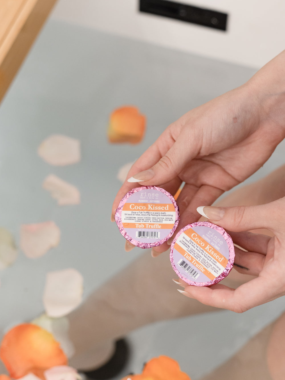 Two hands holding Coco Kissed Tub Truffles above a bath scattered with flower petals — three cocoa butter baths, boxed and ready to give. Rinse Bath & Body Co.