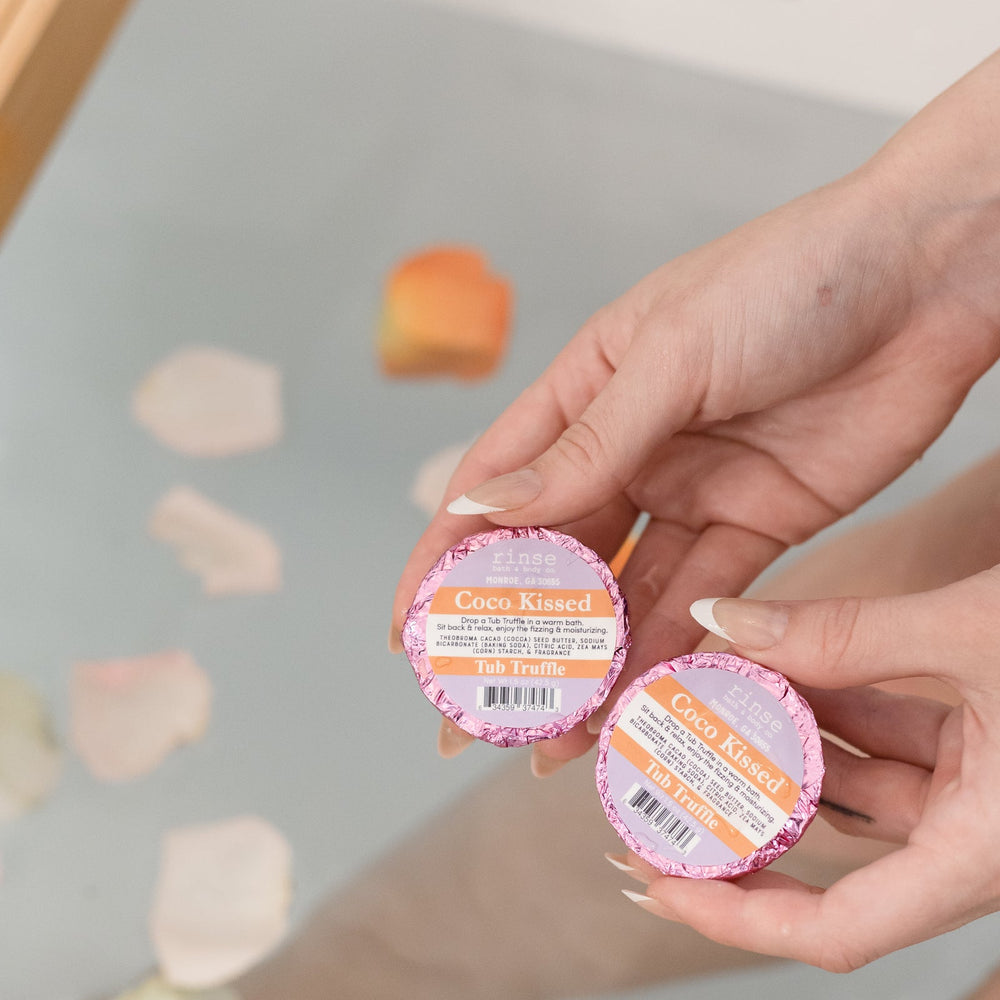 Two hands holding Coco Kissed Tub Truffles above a bath scattered with flower petals — three cocoa butter baths, boxed and ready to give. Rinse Bath & Body Co.