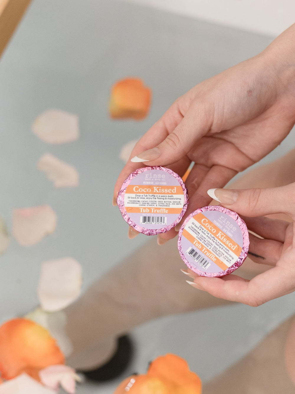 Two hands holding Coco Kissed Tub Truffles above a bath scattered with flower petals — three cocoa butter baths, boxed and ready to give. Rinse Bath & Body Co.