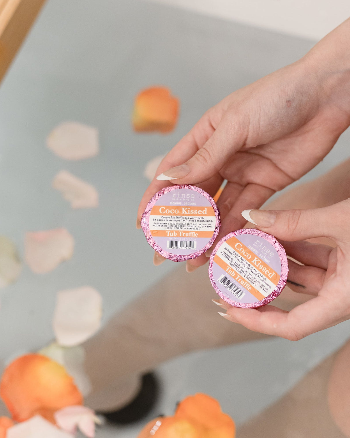 Two hands holding Coco Kissed Tub Truffles above a bath scattered with flower petals — three cocoa butter baths, boxed and ready to give. Rinse Bath & Body Co.